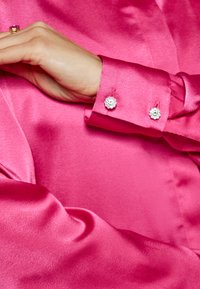 Camicia di raso rosa con una texture liscia, caratterizzata da un polsino decorato con due bottoni circolari instrati di strass.