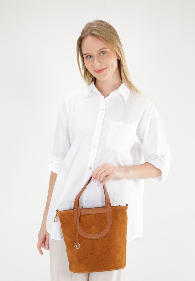 Bruine suede tote bag met leren accenten, ronde handgreep en ritssluiting, vastgehouden door een persoon in een wit shirt tegen een effen achtergrond.