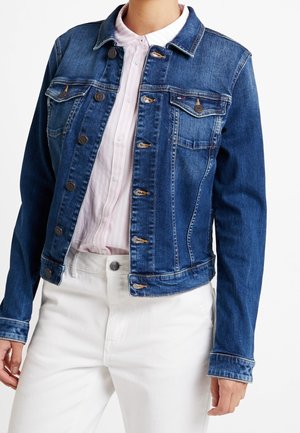 Osoba w niebieskiej dżinsowej kurtce zapinanej na guziki, na jasnoróżowej koszuli zapinanej na guziki i białych spodniach, widziana od szyi do bioder.