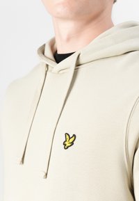 Hoodie beige avec une poche kangourou à l'avant, capuche avec cordon de serrage, et un logo d'oiseau jaune bordé de noir sur la poitrine gauche. Tissu doux et texturé.