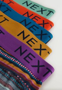 Cinq ceintures colorées pliées avec le mot « NEXT » en lettres majuscules noires sur chacune, disposées en diagonale sur un fond blanc.