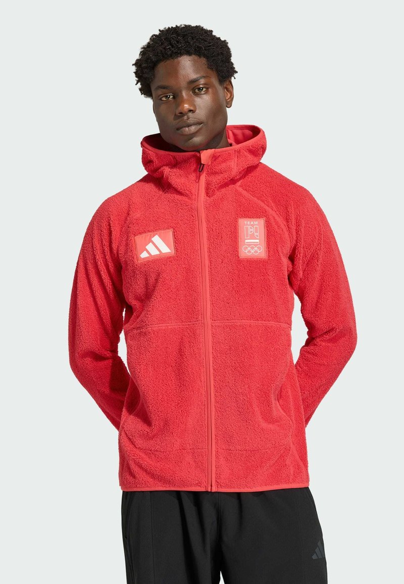 Mężczyzna ubrany w czerwony polar z kapturem, zamkiem z przodu, logo Adidas na lewej piersi oraz logo drużyny olimpijskiej na prawej piersi, z rękami za plecami.