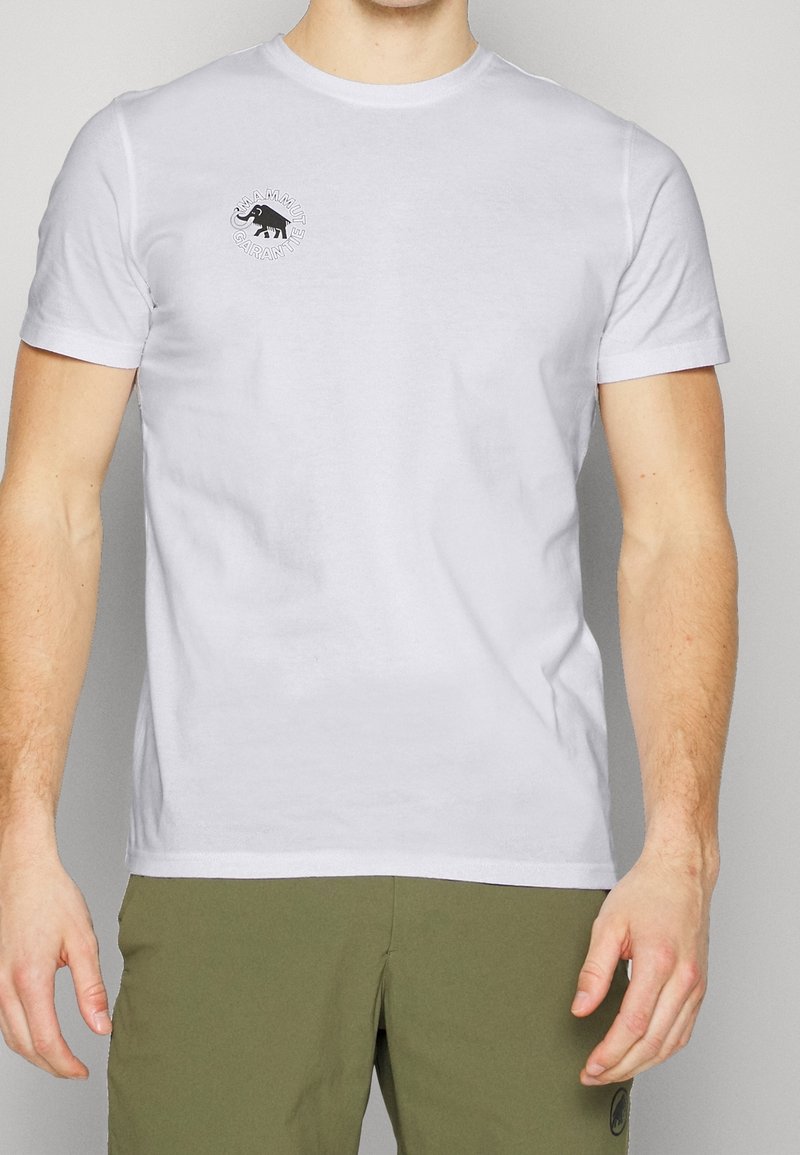 Mann trägt ein schlichtes weißes Kurzarm-T-Shirt mit einem kleinen schwarzen Mammut-Logo und Text auf der linken Brust, kombiniert mit olivgrüner Hose.