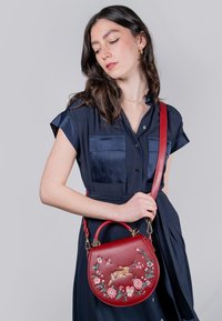 Borsa a tracolla rossa in pelle di forma arrotondata, con ricami floreali e una chiusura in metallo. Cinturino regolabile, texture liscia e colori vivaci.