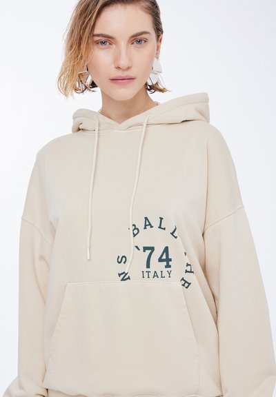 Beige hettegenser med kengurulomme, med grønn trykt tekst "BALL '74 ITALY" i et sirkulært design. Myk, teksturert stoff, justerbar hette.