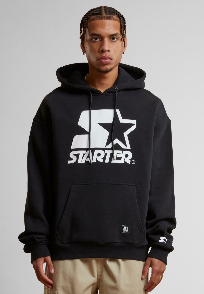 Crna dukserica s velikim bijelim logotipom "STARTER" i dizajnom zvijezde. Model stoji s vidljivim rukama, prikazujući detalje manžeta i kenguru džepa.