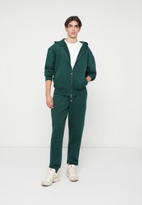 Junger Mann steht in einem dunkelgrünen Reißverschluss-Hoodie, passenden Jogginghosen, einem weißen T-Shirt und beigen Sneakers vor einem weißen Hintergrund.