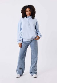 Sweat à zip bleu clair avec poche avant, coupe décontractée et poignets côtelés, associé à un jean en denim clair taille haute et des baskets blanches.