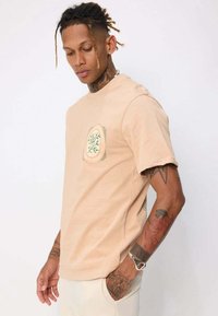 T-shirt en coton beige clair avec un col rond, des manches courtes et un écusson graphique d'arbre sur la poitrine gauche. Offre une coupe décontractée et une texture douce.