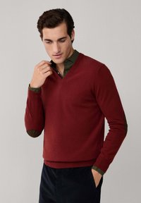 Maglione rosso con scollo a V, texture lavorata a maglia e toppe scure verdi a contrasto sui gomiti. Abbinato a una camicia verde con colletto sotto.