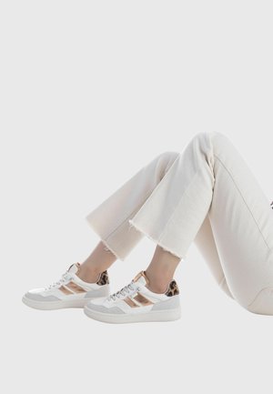 Zapatillas blancas con talón de estampado de leopardo, detalles metálicos y detalle en ante gris. Llevadas con pantalones acampanados color crema, mostradas desde una posición sentada.