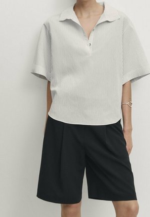 Persona che indossa una camicia bianca larga a maniche corte con sottili righe verticali e pantaloncini neri fino al ginocchio su uno sfondo semplice.