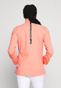 Veste de performance pêche avec manches longues, col montant et bande logo centrale au dos. Texture lisse et design minimaliste.