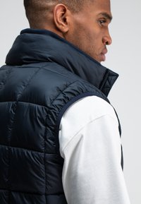 Dunkelblaues gepolstertes Westchen mit hohem Kragen und Reißverschluss, das über einem weißen Hemd getragen wird. Glatte Textur, elegantes Design.