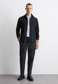Zwarte overhemd met knoop, gedragen over een wit t-shirt. Gecombineerd met zwarte broek en stevige zwarte schoenen, tegen een neutrale achtergrond.