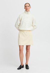 Pull en maille torsadée crème avec un col haut, associé à une mini-jupe en velours côtelé beige clair et des mocassins noirs. Design minimaliste, tissu texturé.