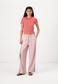 Chemise à rayures rouges et blanches à manches courtes, pantalon large rose, et sandales noires. Le mannequin tient un petit sac à main blanc.