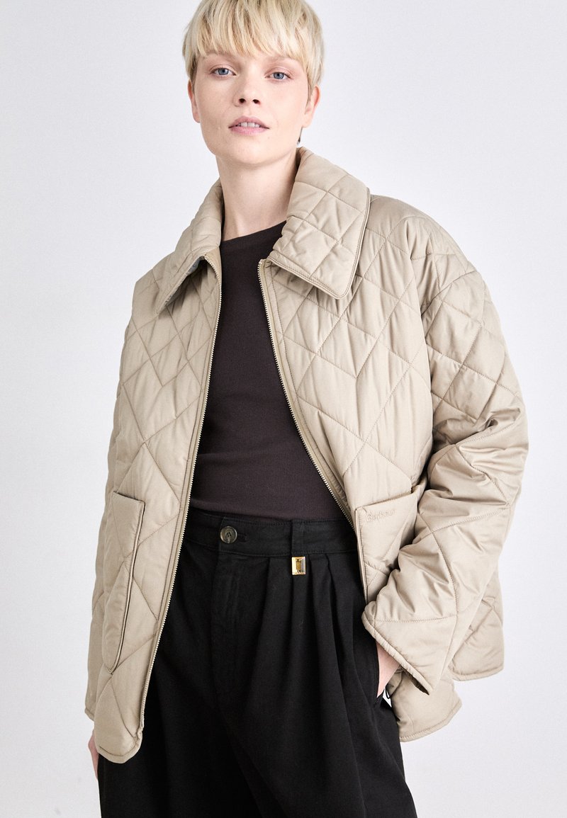 Beige gesteppte Jacke mit großem spitzem Kragen, Frontreißverschluss und schrägen Taschen, kombiniert mit einem dunklen Oberteil und hochtaillierten schwarzen Hosen.