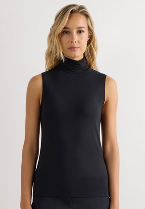 Blonde vrouw die een mouwloze zwarte coltrui draagt, staand tegen een effen witte achtergrond, kijkt recht vooruit.