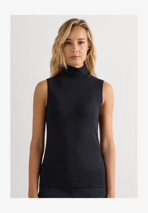 Blond kvinde iført en ærmeløs sort højhalset top, står mod en ensfarvet hvid baggrund og kigger fremad.