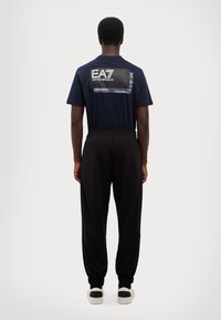 Navy short-sleeve shirt with a large EA7 logo on the back, paired with black tapered joggers featuring elastic cuffs and side pockets.