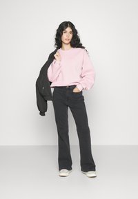 Rosa overdimensjonert sweatshirt med senkede skuldre, kombinert med svarte utsvingte jeans. Svart bomberjakke ligger over den ene armen. Hvite joggesko.