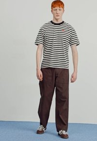 T-shirt à manches courtes noir et blanc à rayures, pantalon large marron, et baskets marron avec des accents blancs, se tenant sur une surface bleue.