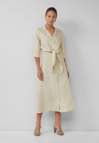 Beige linnenshirt met driekwart mouwen, knoopsluiting aan de voorkant en een strik in de taille, gecombineerd met een bijpassende maxi rok en hoge sandalen.