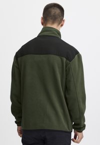 Grønn fleecejakke med svart skulderstykke, høy krage og lange ermer. Myk tekstur med avslappet passform. Ingen synlige mønstre eller detaljer.