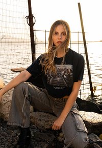 Zwarte cropped t-shirt met een grafische print, gecombineerd met losse grijze cargobroeken. De setting omvat stenen en water, verlicht door de zonsondergang.