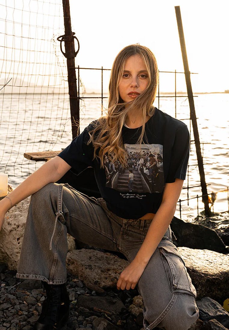 Zwarte cropped t-shirt met een grafische print, gecombineerd met losse grijze cargobroeken. De setting omvat stenen en water, verlicht door de zonsondergang.