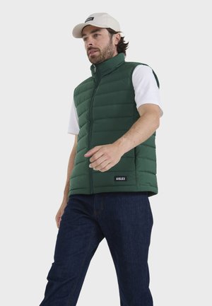 Chaleco acolchado verde con cremallera en la parte delantera, textura acolchada y cuello alto, combinado con una camiseta blanca y jeans azules. Logo de la marca visible.