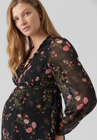 Vestido de maternidad de tejido negro transparente con estampado floral en rosa, naranja y verde. Presenta mangas largas y un diseño de cintura fruncida.