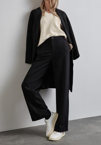 Pantalon noir sur mesure associé à un pull à côtes crème et à un long manteau noir. Des baskets blanches complètent la tenue. Design simple et épuré.