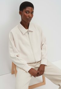 Donna con capelli corti che indossa una giacca beige chiaro con zip e pantaloni, seduta su uno sgabello di legno su sfondo bianco.