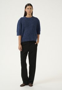 Maglione blu lavorato a maglia con maniche corte, texture a coste, abbinato a pantaloni neri e scarpe nere intrecciate, su uno sfondo chiaro.