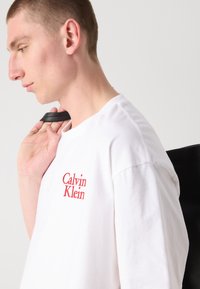 Balta medvilnė marškinėliai su raudona išsiuvinėta "Calvin Klein" logotipu kairiajame krūtinės kampe, dėvimi su juodu diržu per petį.