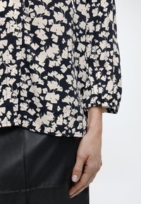 Μπλούζα μακρυμάνικη με ασπρόμαυρα λουλούδια και κουμπιά, σε συνδυασμό με μαύρη δερμάτινη φούστα, εμφανιζόμενη από τη μέση έως το χέρι στη δεξιά πλευρά.