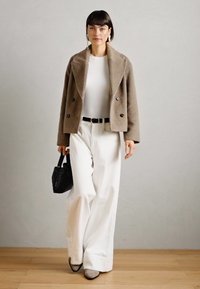 Veste en laine beige à double boutonnage, haut blanc ajusté, large pantalon blanc, ceinture noire, petit sac à main noir et chaussures grises.