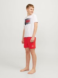 Maglietta bianca a maniche corte con design grafico nero e rosso; pantaloni da bagno rossi con logo; piedi nudi. Abbigliamento semplice e informale.