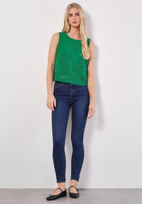 Grünes ärmelloses Oberteil mit Spitzenverzierungen, kombiniert mit dunkelblauen engen Jeans und schwarzen Ballerinas. Einfaches, lässiges Outfit.