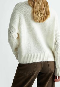 Žena s rameny dlouhými blond vlasy, oblečená v bílém pleteném svetr s copánkovými detaily na rukávech a hnědých kalhotách s vysokým pasem, obrácená zády.