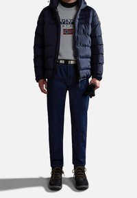 Giacca puffer blu scuro su una maglietta grigia con grafica norvegese, jeans blu e scarponi da trekking neri con dettagli dorati.