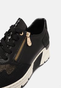 Svart sneaker med blandade texturer, med en glansig lackad ovandel, gulddragkedja och en kraftig vit sula med svarta accenter.