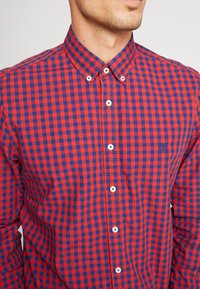 Camicia a quadri rossa e blu con colletto button-down, dotata di bottoni bianchi e logo sobrio sul petto. Tessuto in cotone, texture liscia.