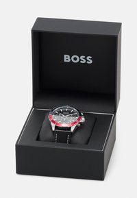 Reloj BOSS en caja negra; presenta caja metálica plateada, esfera negra, bisel rojo, correa de cuero negro con costuras blancas y detalle de cronógrafo.
