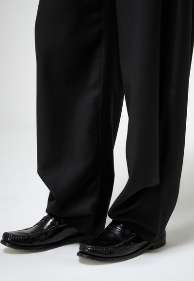 Juodos platėjančios kelnės iš lygaus audinio, derinamos su blizgančiais juodais loaferiais, turinčiais siuvinėtą dizainą ir nedidelį kulniuką.