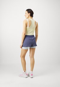 Αθλητική εμφάνιση που περιλαμβάνει ένα ανοιχτό κίτρινο μπλουζάκι με ανοιχτή πλάτη, συνδυασμένο με ναυτικές βερμούδες και λευκά αθλητικά παπούτσια με ροζ λεπτομέρειες.