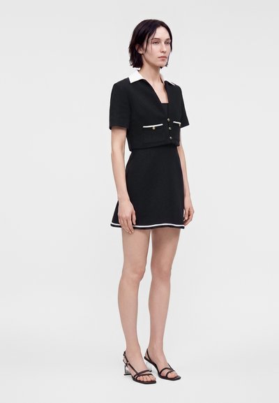 Femme debout portant une robe noire à manches courtes avec une bordure blanche, des poches avant, un col blanc et des sandales noires à talons sur fond blanc.