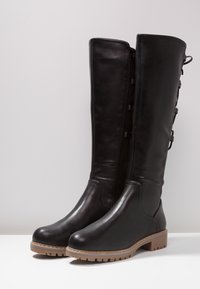 Pár černých kožených holínek až po kolena se bočním zipem, tkaničkovou detailní úpravou na zadní straně a světle hnědými gumovými podrážkami, vystavených na bílé ploše.